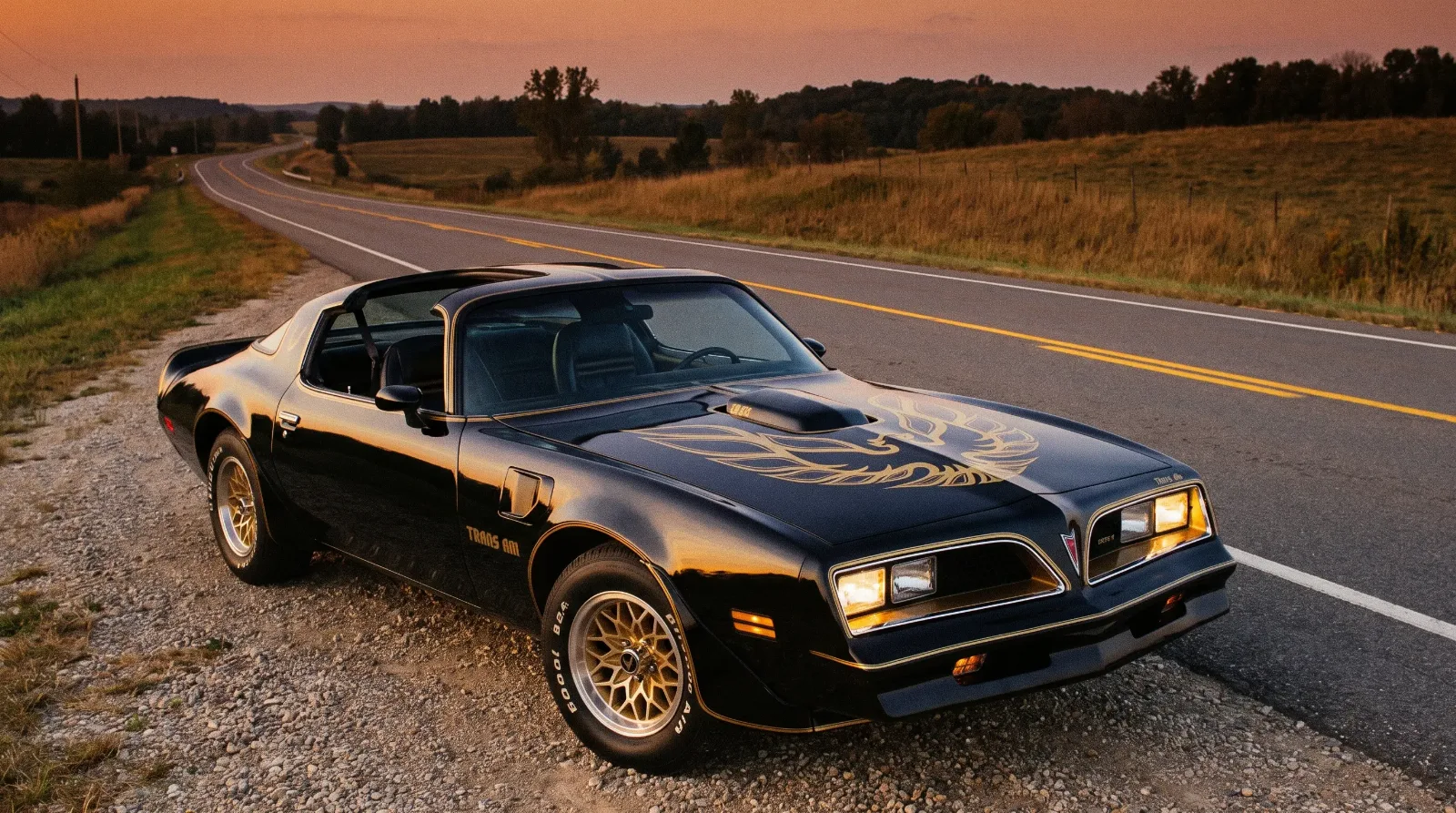 Black and gold 1977 Pontiac Trans Am with T-tops off parked on a rural two-lane highway at dusk