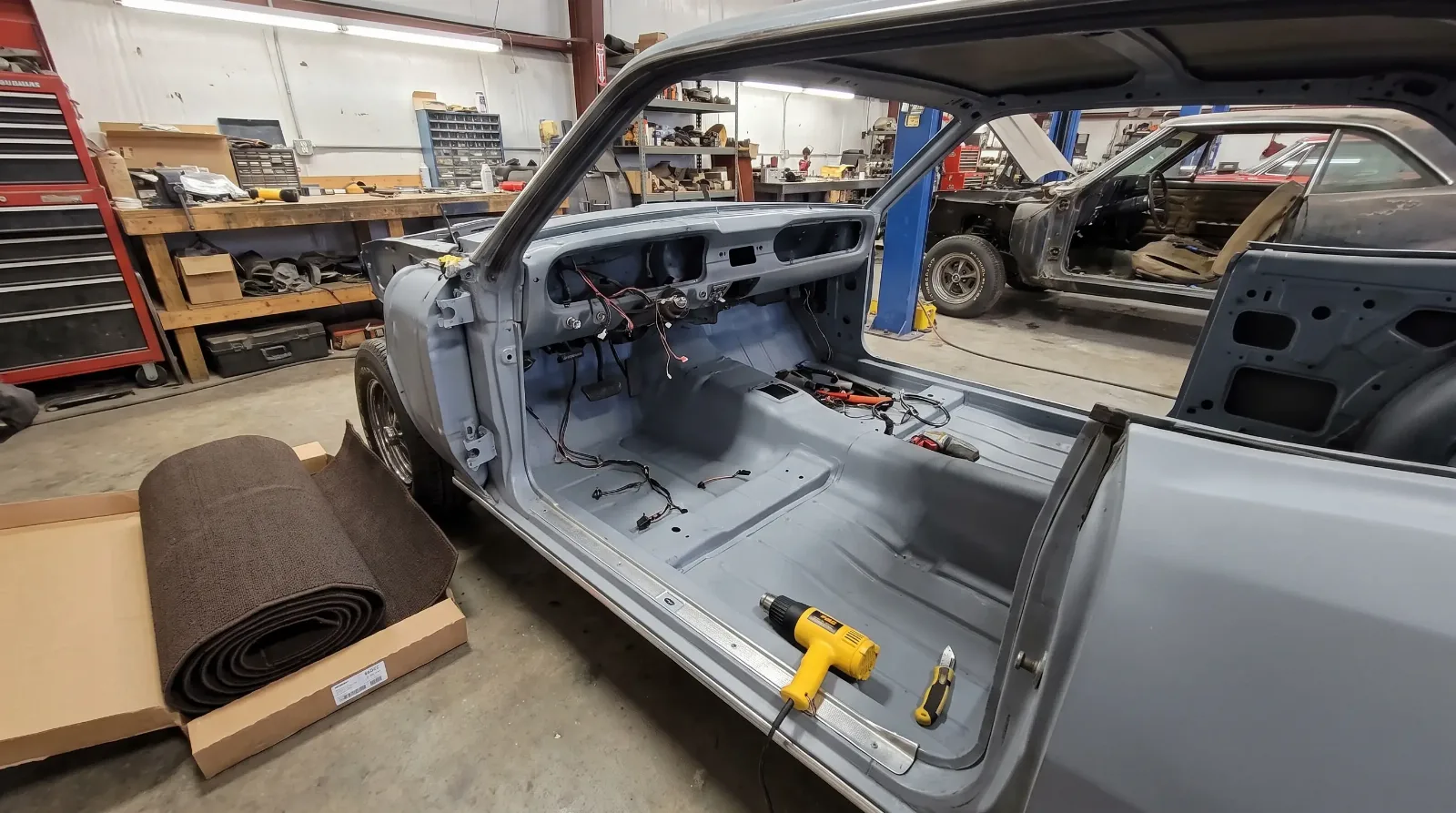 Primed floor pan of a 1960s coupe with a new molded carpet kit laid out beside the car in a restoration shop