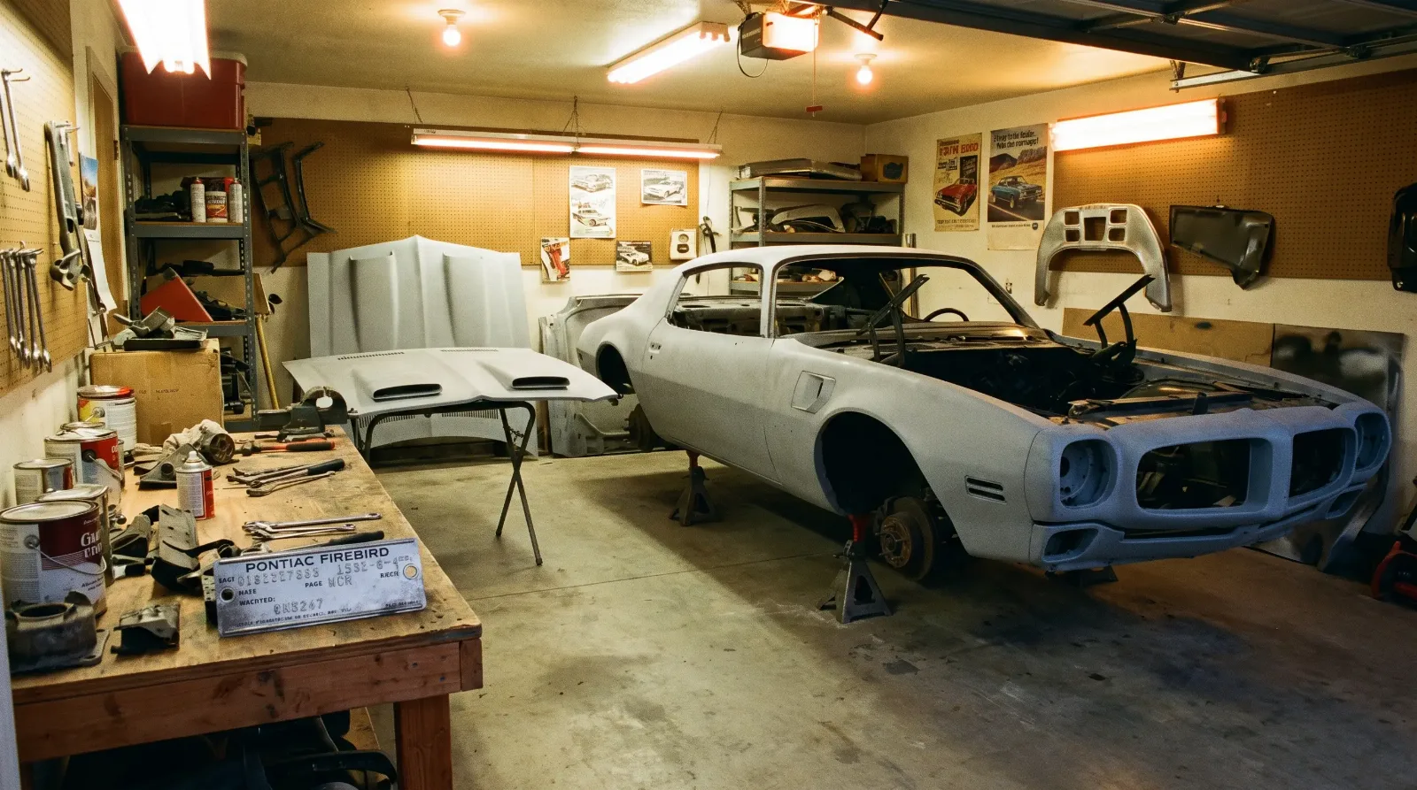 Second-generation Pontiac Firebird body shell on jack stands in a garage with a Formula twin-scoop hood test-fitted nearby