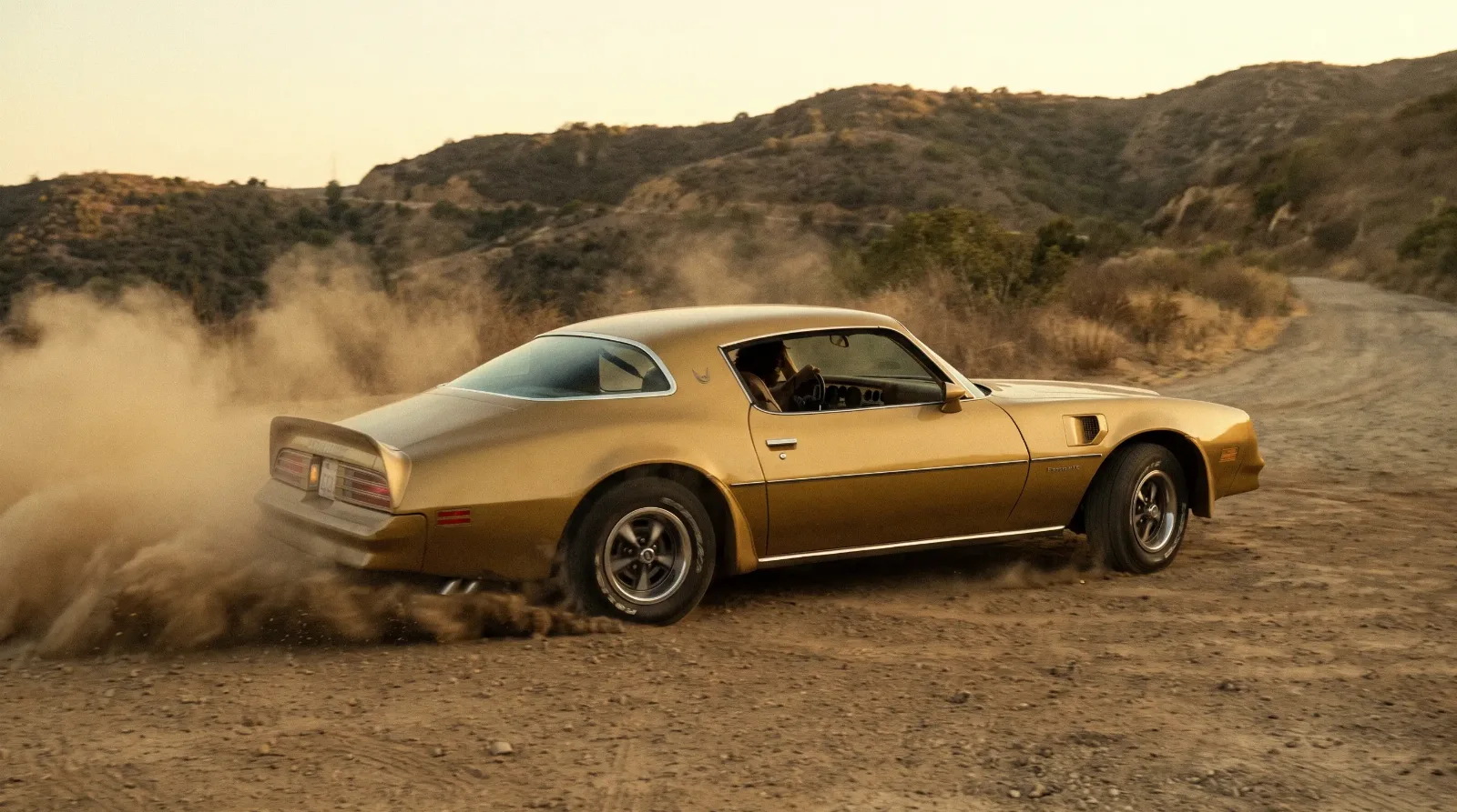 Gold Pontiac Firebird Esprit performing a reverse J-turn on a dusty dirt road in 1970s style
