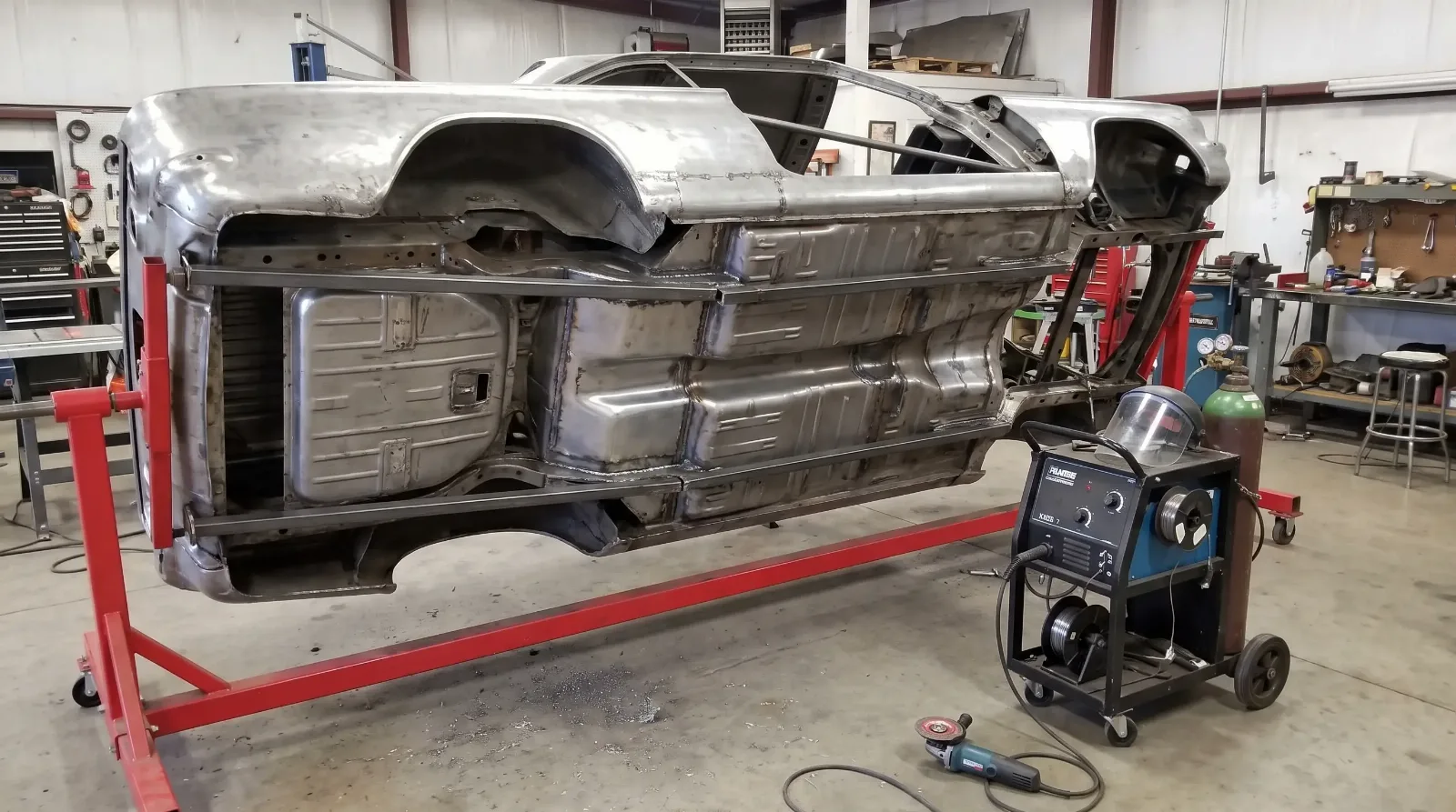 Bare-metal unibody shell of a 1960s coupe turned on its side on a rotisserie, with bracing welded across the door openings and new floor patches