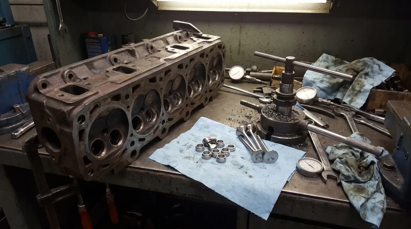 Bare cast-iron V8 cylinder head on a machine shop bench with hardened valve seat inserts and valves laid out beside it