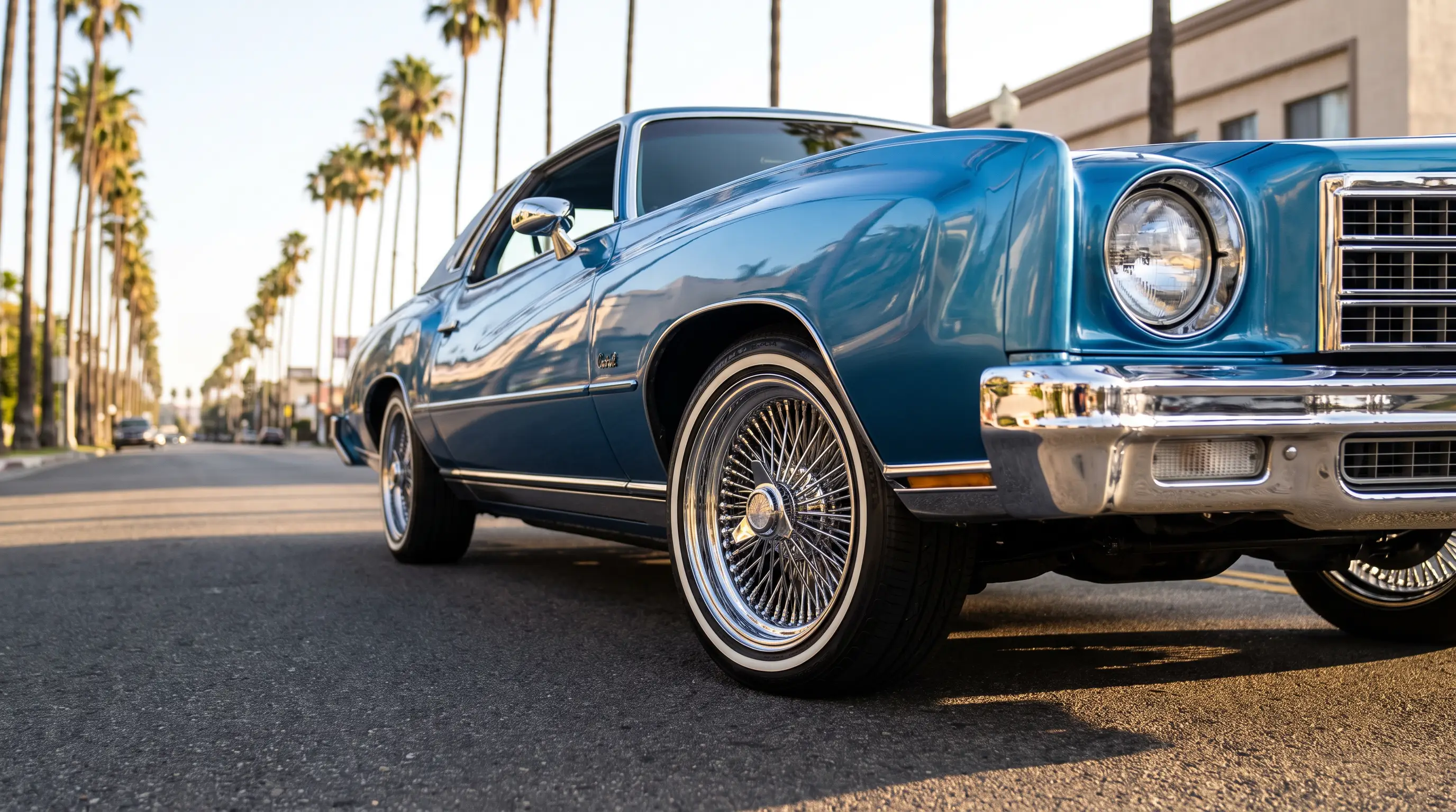 Lowrider Chevrolet Monte Carlo with polished wire wheels and whitewalls on a sunlit boulevard