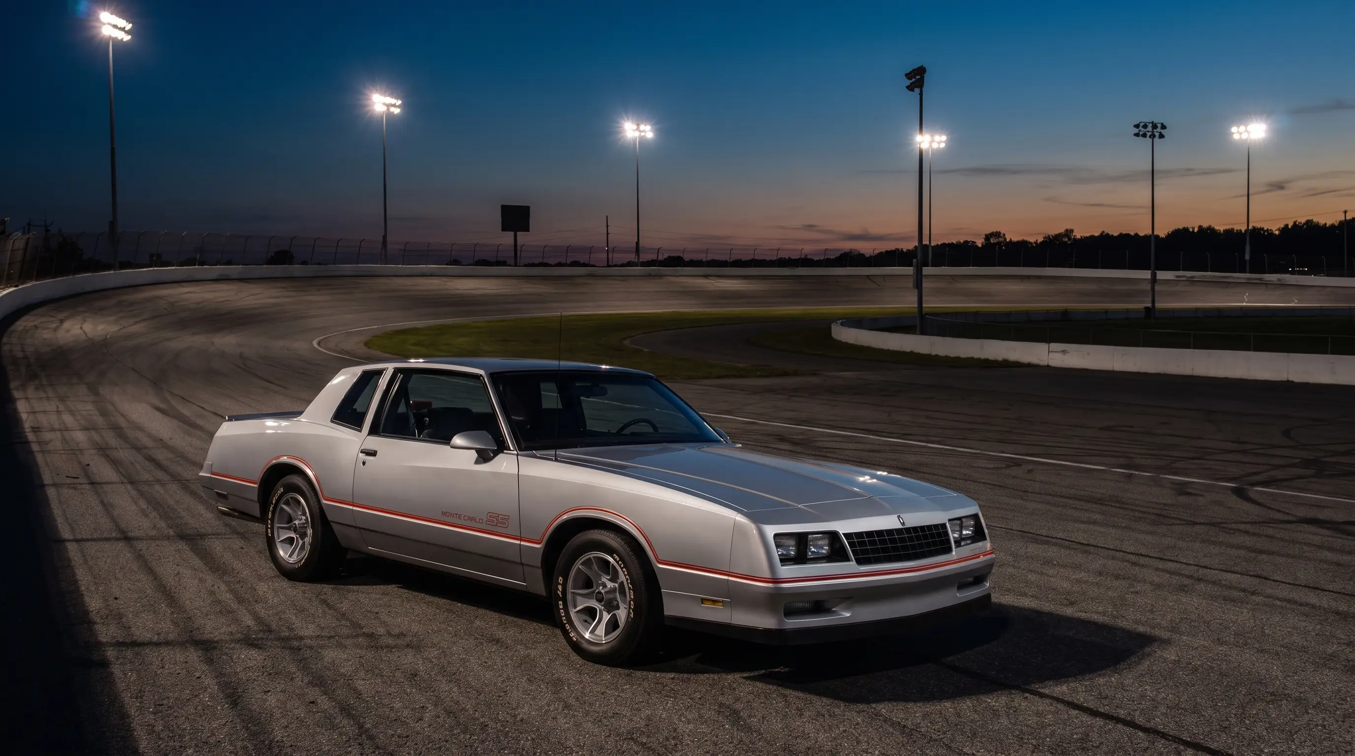 Silver 1983 Chevrolet Monte Carlo SS beside a banked NASCAR oval at dusk