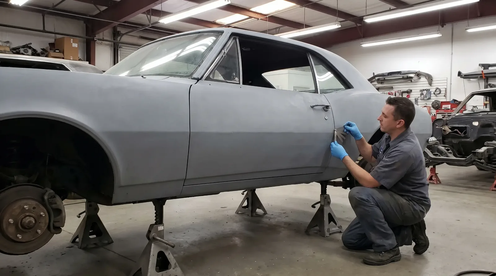 Restoration technician checking a door gap with a feeler gauge on a 1960s coupe in gray primer