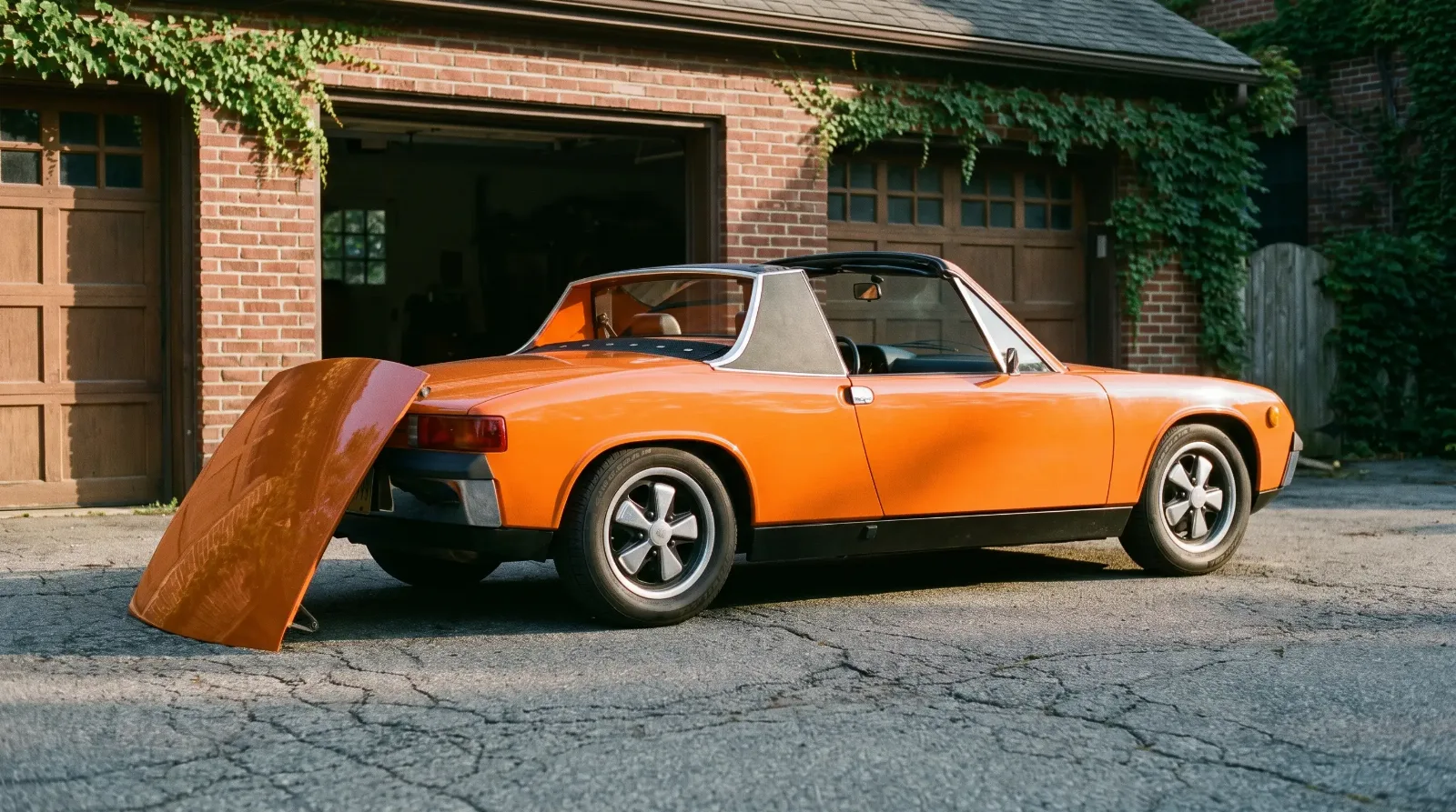 1973 Porsche 914 in Signal Orange with targa roof panel removed, parked outside a brick garage at golden hour