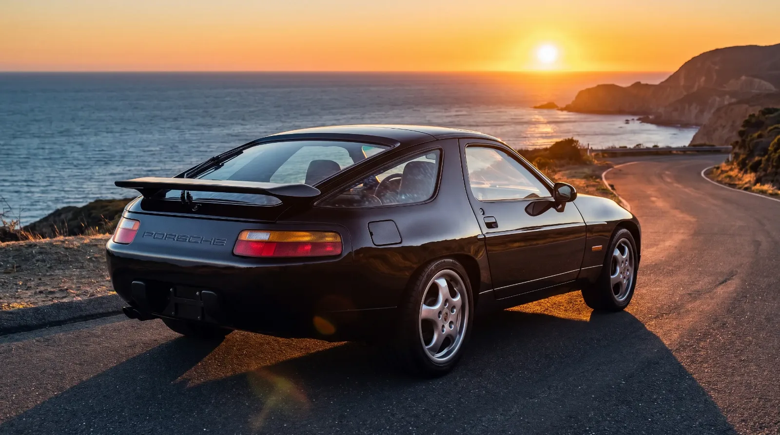 Black Porsche 928 GTS on a coastal road at golden hour showing its widened rear fenders