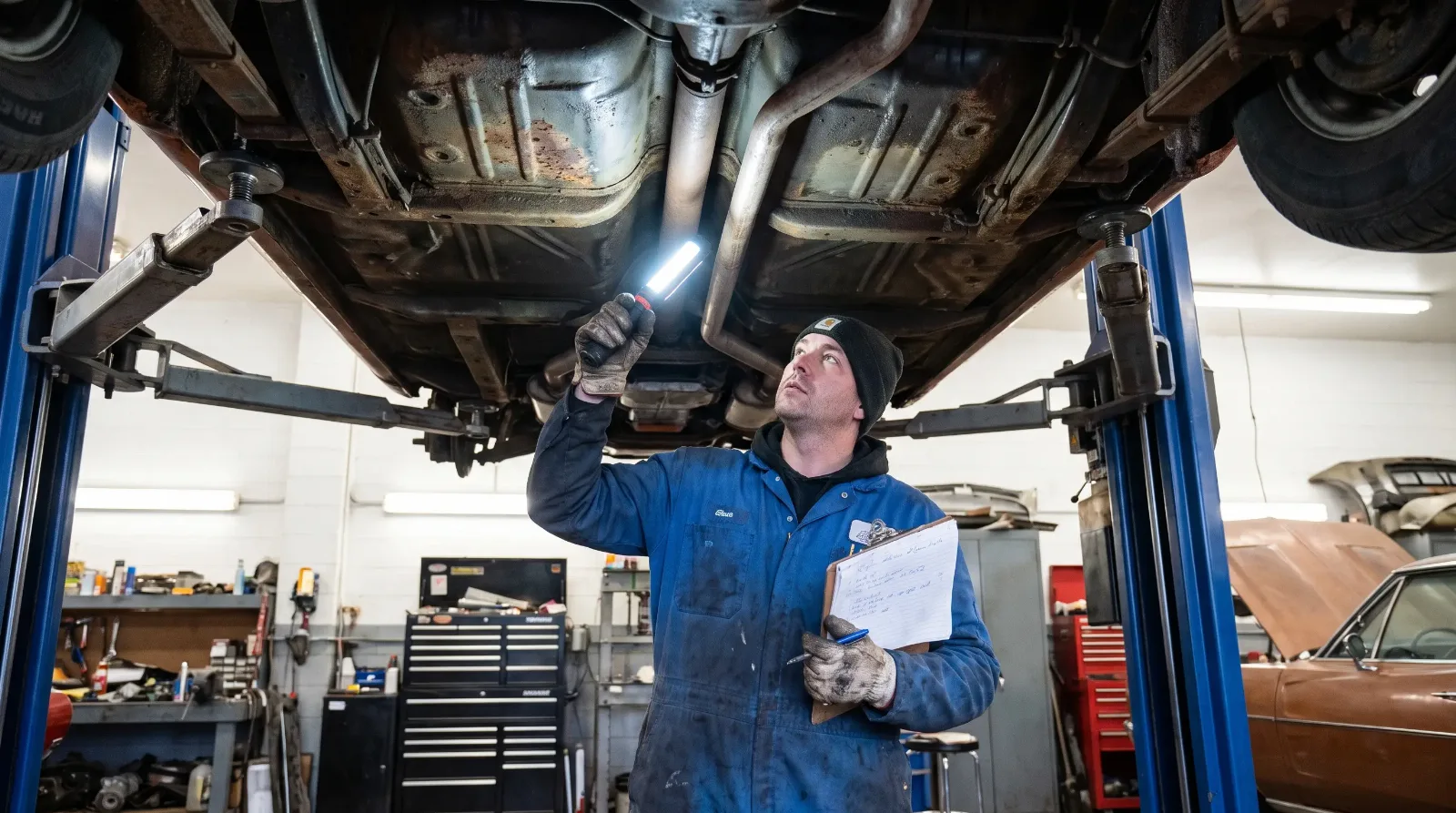 Mechanic inspecting the frame rails of a classic muscle car raised on a two-post lift with a flashlight
