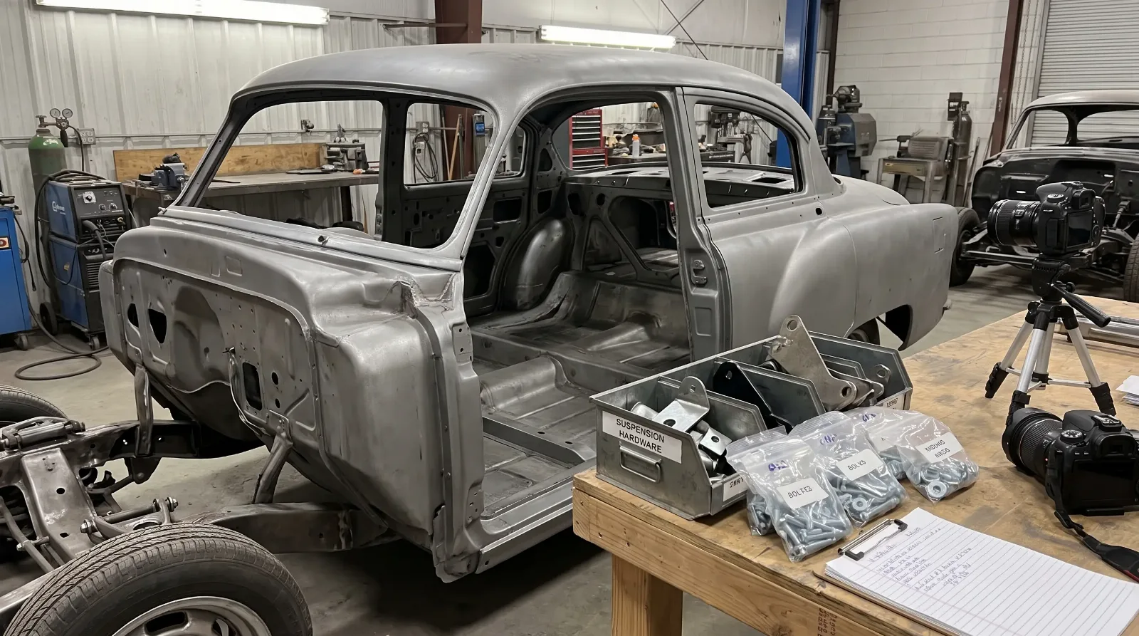 Media-blasted bare-metal sedan shell in a restoration shop with bagged and tagged parts on a bench and a camera on a tripod